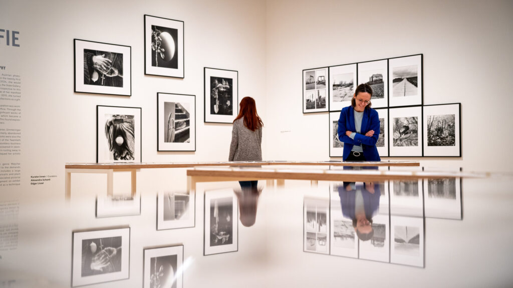 Man sieht zwei Frauen, die in der Ausstellung der Landesgalerie von Elfriede Mejchar stehen und die Fotografien betrachten, 