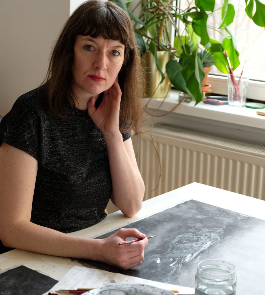 Alexandra Kontriner in ihrem Atelier beim Arbeiten. Hinter ihrem Schreibtisch auf dem Fesnterbrett sieht man eine grüne Pflanze.